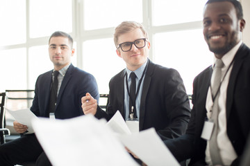 Adult multiracial men in suits sitting with papers in hands and communicating on business meeting