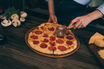 Woman cutting fresh baked homemade pizza on rustic kitchen background. Cut into slices delicious pizza with mushrooms and ham. Cheese and tomatoes on wooden table. Healthy foods, cooking concept.
