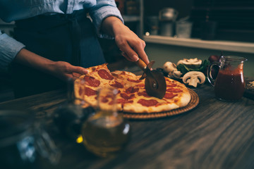 Woman cutting fresh baked homemade pizza on rustic kitchen background. Cut into slices delicious pizza with mushrooms and ham. Cheese and tomatoes on wooden table. Healthy foods, cooking concept.
