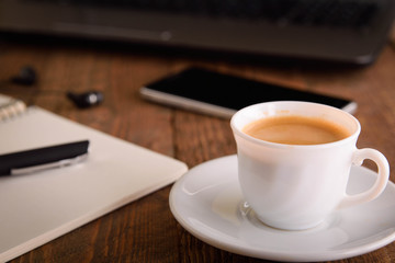 Cup of coffee on a laptop keyboard. Work place modern. Laptop (notebook) with cup of coffee and notepad with pen on old wooden table.