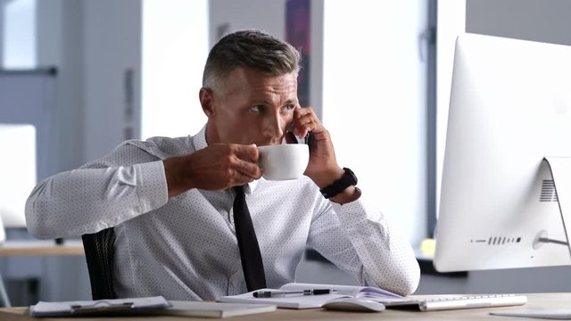 Serious concentrated businessman in white shirt talking on phone and drinking coffee while working in office