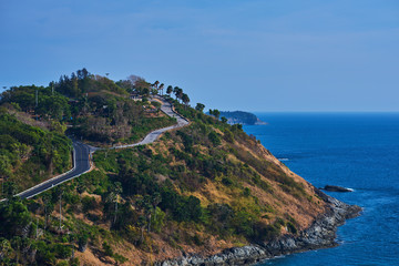 View on the rocky coastline with green tropical trees, yellow grass and asphalt coastal road going round edge bend on a sunny day with blue sky and ocean. Beautiful nature landscape.