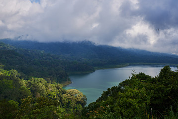Fototapeta premium Forested mountain slope in low lying cloud with the evergreen tropical trees, shrouded in mist in a scenic landscape. Low cloud shrouds the distant mountains above the waters of the lake.