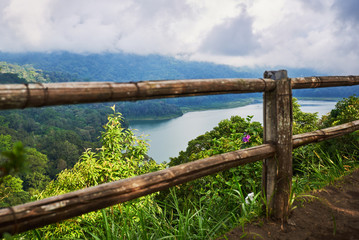 Naklejka premium Wooden fence along the shore of the lake. Low cloud shrouds the distant mountains. Tropical landscape with dramatic clouds in the sky. Lake and mountain view from a hill. Nature background.