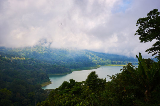 Forested mountain slope in low lying cloud with the evergreen tropical trees, shrouded in mist in a scenic landscape. Low cloud shrouds the distant mountains above the waters of the lake.