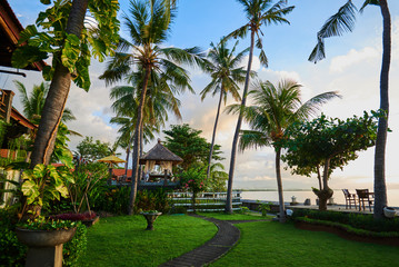 Fototapeta premium Beautiful view on the tropical bungalow in the green garden with palms and flowers. Asian beach resort. Exotic landscaping design. Stone footpath to paradise beach. Vacation and travel concept.