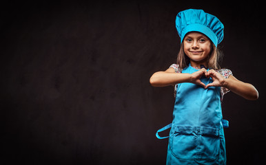Happy little girl dressed in blue cook uniform shows love gesture. Isolated on a dark textured...