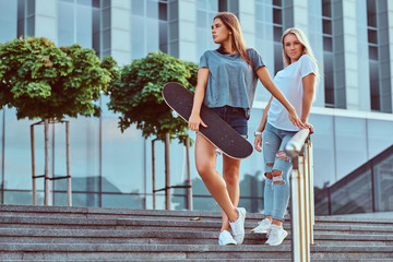 Two beautiful hipster girls standing on steps with skateboard on a background of the skyscraper. © Fxquadro