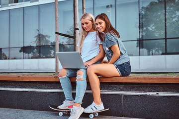 Two beautiful hipster girls sitting on the bench with a laptop on a background of the skyscraper. © Fxquadro