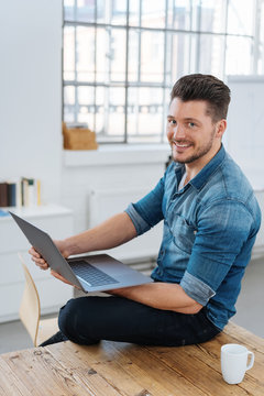 lächelnder junger mann sitzt auf dem tisch mit seinem laptop 