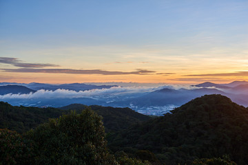 Mountain hill covered with forest on foreground and curly clouds on background with foggy valley with village in between. Mountains during sunrise. Beautiful natural landscape in the summer time..