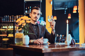 Portrait of a handsome man with stylish beard and hair dressed in a black suit sitting at the cafe, holds a glass of wine.