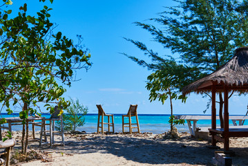 Beach bar with bamboo chairs on sand and thatched roof tropical hut on the shining turquoise ocean background. Beach bar view towards the ocean. Relaxing atmosphere. Travel and holiday concept.