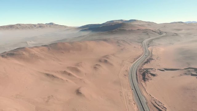 Aerial cinematic view of highway road with traffic, rocky sand dune with mist and fog. Atacama desert Iquique Chile. Drone flying backward at beautiful sunrise.