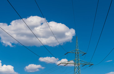 Transmission tower top part on a blue sky background. Wires of power line on electricity pylon detail against beautiful heaven with white fluffy clouds. Sunny summer day. Idea of industry, ecology.