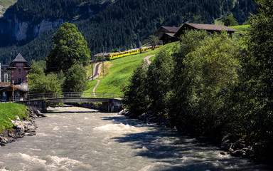Fototapeta na wymiar Picturesque alpine scene with a river, some typical wooden chalets and a yellow train coming down the mountain. Grindelwald, Bernese Oberland, Switzerland