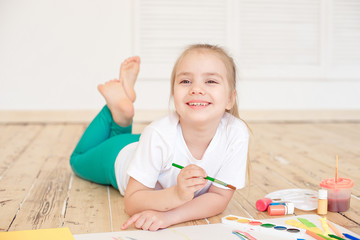 Little blonde girl painting on big white paper while laying on the floor indoors.
