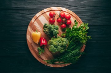 Fresh vegetables: cherry tomatoes, broccoli, pepper, dill, parsley on a round wooden board, on a dark wooden table. Recipe. Ingredients. Dietary food. Place under the text. View from above