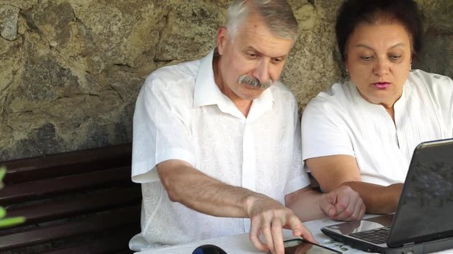 mature senior couple do online shopping on computer. both in white shirt staring at laptop pointing at computer screen. man take smartphone ready to make order. static close side outdoor shot