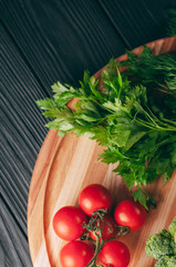 On a dark wooden table, on a round wooden board fresh green broccoli, parsley, dill and cherry tomatoes for your health. Recipe. Ingredients. Dietary food. Place under the text. View from above