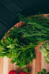 On a dark wooden table, on a round wooden board fresh green broccoli, parsley, dill and cherry tomatoes for your health. Recipe. Ingredients. Dietary food. Place under the text. View from above