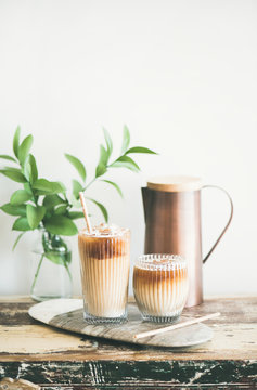 Iced coffee in glasses with milk and straws on board over rustic wooden table, white wall, jug and plant branch in vase at background, copy space. Summer refreshing beverage, ice coffee drink concept