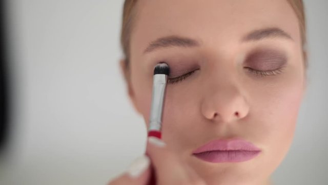 Professional make-up artist visagiste blending dark brown eye shadows with brush sponge on eyelid. Woman client in beauty salon doing makeup close up indoors slow motion. Facial cosmetics care