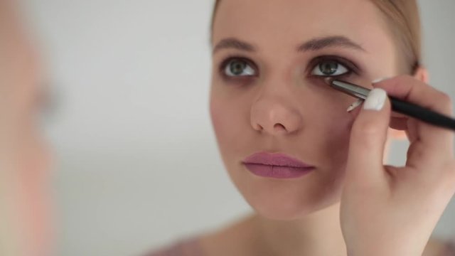 Professional make-up artist applying eyeliner on female eyelid with black pencil in beauty salon. Smiling girl models face detail facial cosmetics close up indoors slow motion. Fashion industry skin