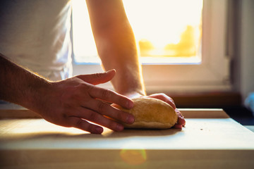 The man, the cook holds a bread roll in his hands, the bread on the counter. The man is holding bread in the hands against the background of the setting sun in the kitchen.