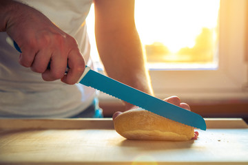The man, the chef cuts a bread roll with a kitchen knife, the bread lying on the counter. The man is holding bread in the hands against the background of the setting sun in the kitchen.