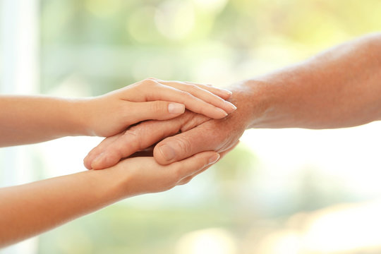 Young woman holding elderly man hand on blurred background, closeup. Help service
