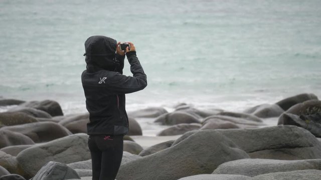 A woman takes pictures of the cold turquoise sea in Unstad as she stands on the rocky beach near the edge of the water. Her jacket with hood pulled up keeps her warm and dry. Slow motion.