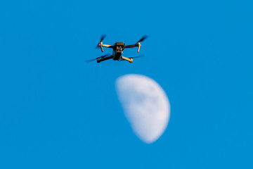 A consumer quadcopter camera drone hovering with a blue sky and the moon behind