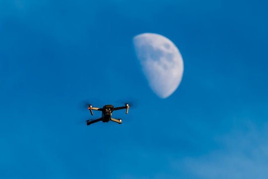A consumer quadcopter camera drone hovering with a blue sky and the moon behind