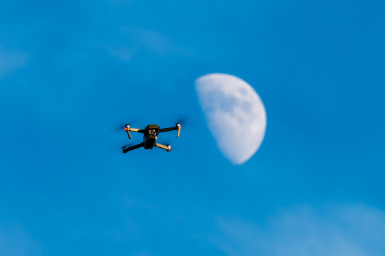 A consumer quadcopter camera drone hovering with a blue sky and the moon behind