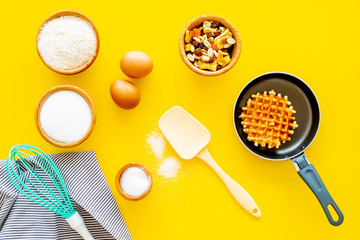 Ingredients for belgian waffles. Eggs, flour, sugar, dried fruits near waffle on pan, whisk for whipping on yellow background top view