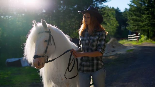 Portrait of a young girl in a cowboy hat near a horse, close-up. Slow motion. Young woman near a horse in evening sunset light.