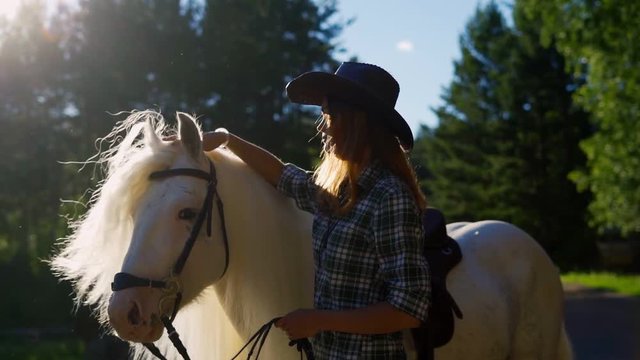A young girl in a cowboy hat takes care of and caresses a horse on an animal farm on a hot summer day. Slow motion