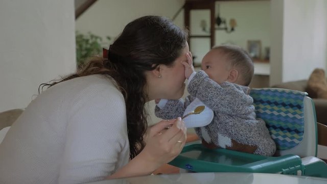 Mother feeding his smiling baby boy with green vegetables pap in living room at home while he tries to bite her nose to play. Winter time clothing. Child routine, day-to-day activities.