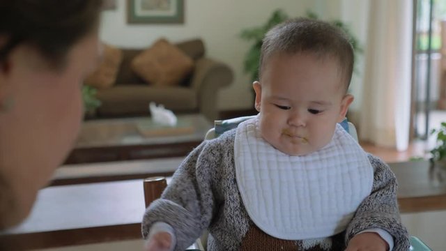 Mother feeding his excited baby boy with green vegetables pap in living room at home. Winter time clothing. Child routine, day-to-day activities.