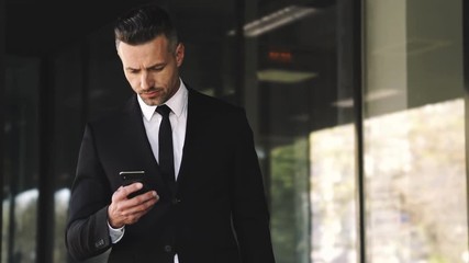 Serious business man dressed in suit using smartphone while waitng someone outside a glass building - Powered by Adobe