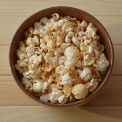 Popcorn in a brown ceramic bowl on a wooden table. Top view, square.