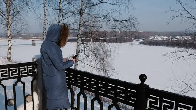 Unrecognizable woman in blue down jacket writes messaging in her cellphone in winter Park.