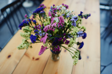 Small vase full of wild field flowers sitting on a tabletop of light wood. Long table for parties, glass vase of with purple flowers on top.