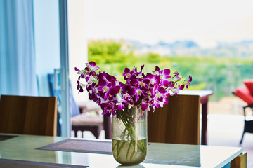 Restaurant, table at a window with a bunch of flowers.Bouquet of violet orchids in glass vase near the window, horizontal photo Selective focus. Elegant violet orchids in glass vase stand on the table