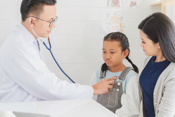 Professional doctor wearing white coat using stethoscope to examine kid patient with her mother in hospital background.Concept of disease treatment and health care in hospitals.