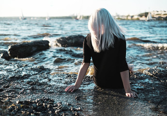 The girl with blond hair sits back on shore of lake sea and looks into the distance at boats