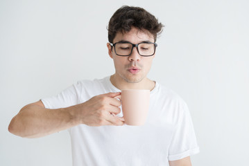 Portrait of young Asian man blowing at hot coffee in cup. Vietnamese student wearing glasses...