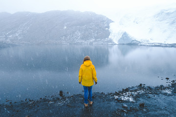 A girl in a yellow jacket and jeans stands on the shore of a blue lake in the snow, on the shore of snowy glaciers