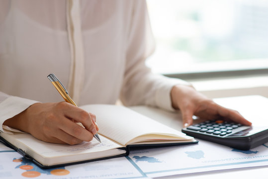 Business expert analyzing reports and counting expenses. Female accountant sitting at table with paper diagrams, writing in notebook and using calculator. Accounting concept 
